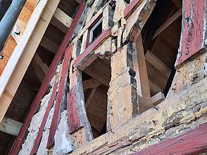 Man sieht einen Ausschnitt einer Fachwerkfassade. Das Holz ist alt und beschädigt. Teilweise wurde das Mauerwerk zwischen den Hölzern entfernt und man sieht Löcher in der Fassade.