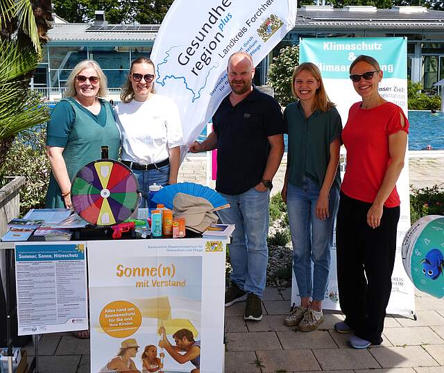 Vier Frauen und ein Mann in der Mitte befinden Sie vor einem Außenschwimmbecken an einem Aktionsstand zum Thema Sonnenschutz. Am Stand befinden sich Banner, Infomaterialien, Sonnencremes und Sonnenkäppis.