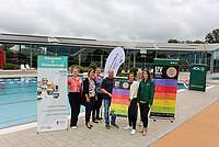 Mehrere Personen vor einem Freischwimmbecken mit Roll-ups und Flaggen, die zum Thema UV-Index erläutern. Im Hintergrund ein Ausschnitt der Glasfront eines Freizeitbades.
