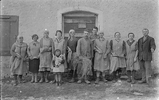 Schwarzweiß-Aufnahme von Personen in ländlicher Umgebung vor einem Haus.