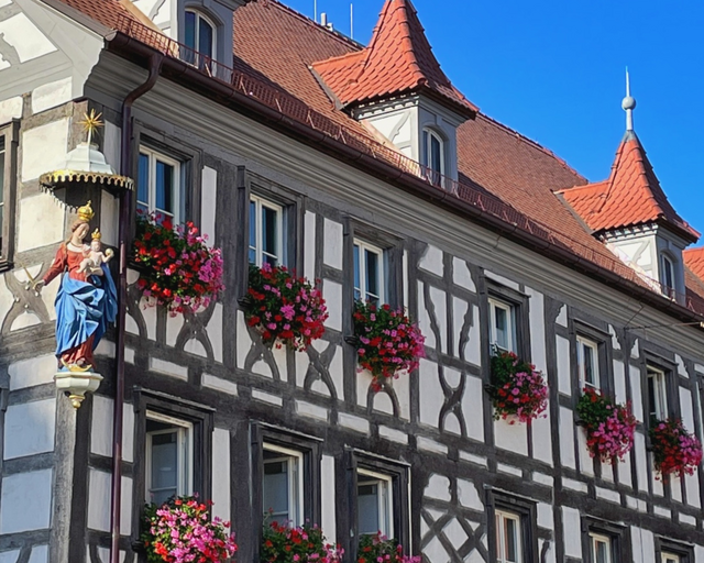 Ein Fachwerkgebäude mit Blumenschmuck an den Fenstern und mit Dacherkern und einer Marienfigur an der Hausecke.