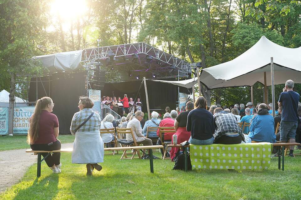 Stadtbadserenade im Königsbad Forchheim im Rahmen des KulturSommerQuartiers Zuschauer, die auf Bierbänken vor einer großen Bühne sitzen und einem Chor zuhören.