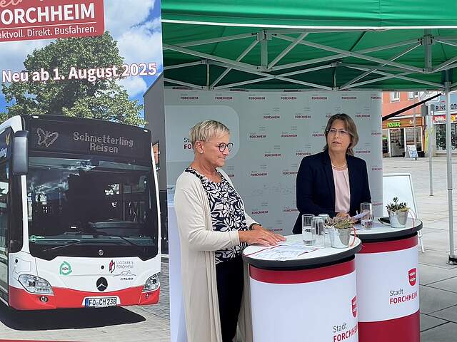 Zwei Frauen stehen an Rednerpulten unter einem Pavillon. Links im Bild ein Rollup mit einem Linienbus.