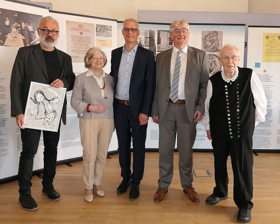 Eine Gruppe aus vier Männern und einer Frau vor mehreren Infowänden mit Fotos, Urkunden und Infotexten. Der Mann links im Bild hält eine Zeichnung in den Händen. Das Motiv zeicgt ein Mädchen mit geflochtenen Zöpfen und ein Haus.