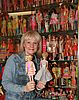 Eine Frau hält zwei Barbie-Puppen in den Händen. Hinter ihr befinden sich mehrere Glasvitrinen mit vielen unterschiedlichen Barbiepuppen.