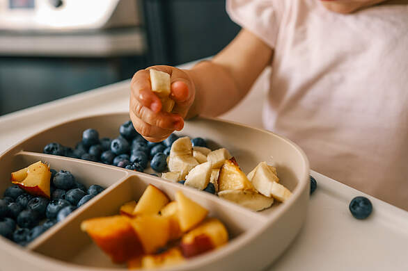 Großaufnahme eines Kindertellers mit geschnittenem Obst und Beeren von dem ein Kleinkind gerade isst.