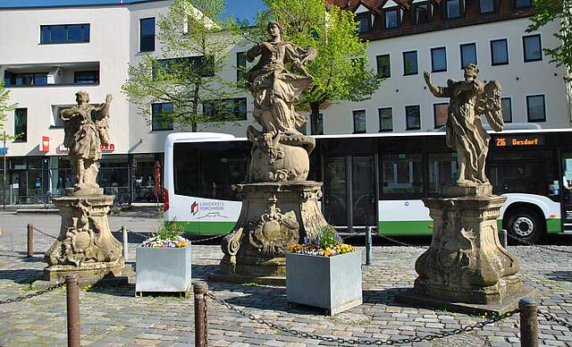 Drei Figuren auf einem Platz, im Hintergrund ist ein Omnibus und eine Häuserfassade zu sehen. 