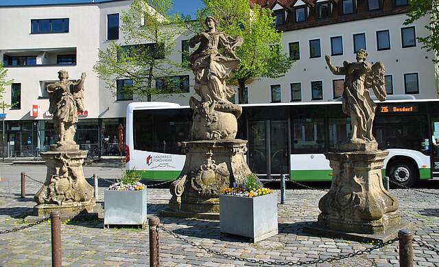Drei Figuren auf einem Platz, im Hintergrund ist ein Omnibus und eine Häuserfassade zu sehen. 