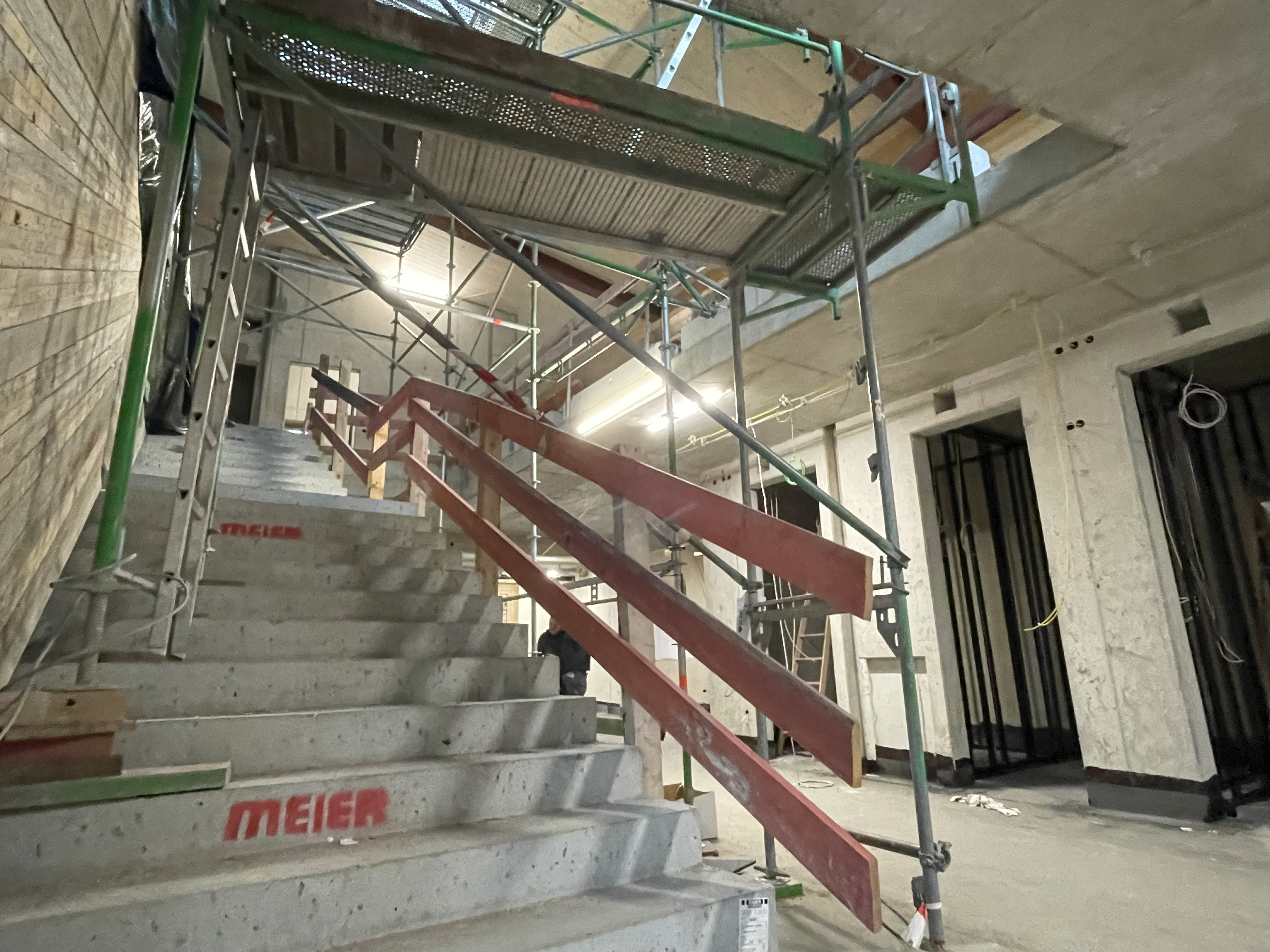 Treppenaufgang zum Obergeschoss. Für die barrierefrei Nutzung wird es einen zusätzlichen Aufzug geben. Blick vom Erdgeschoss nach oben auf eine massive Treppe mit einem Baugerüst.