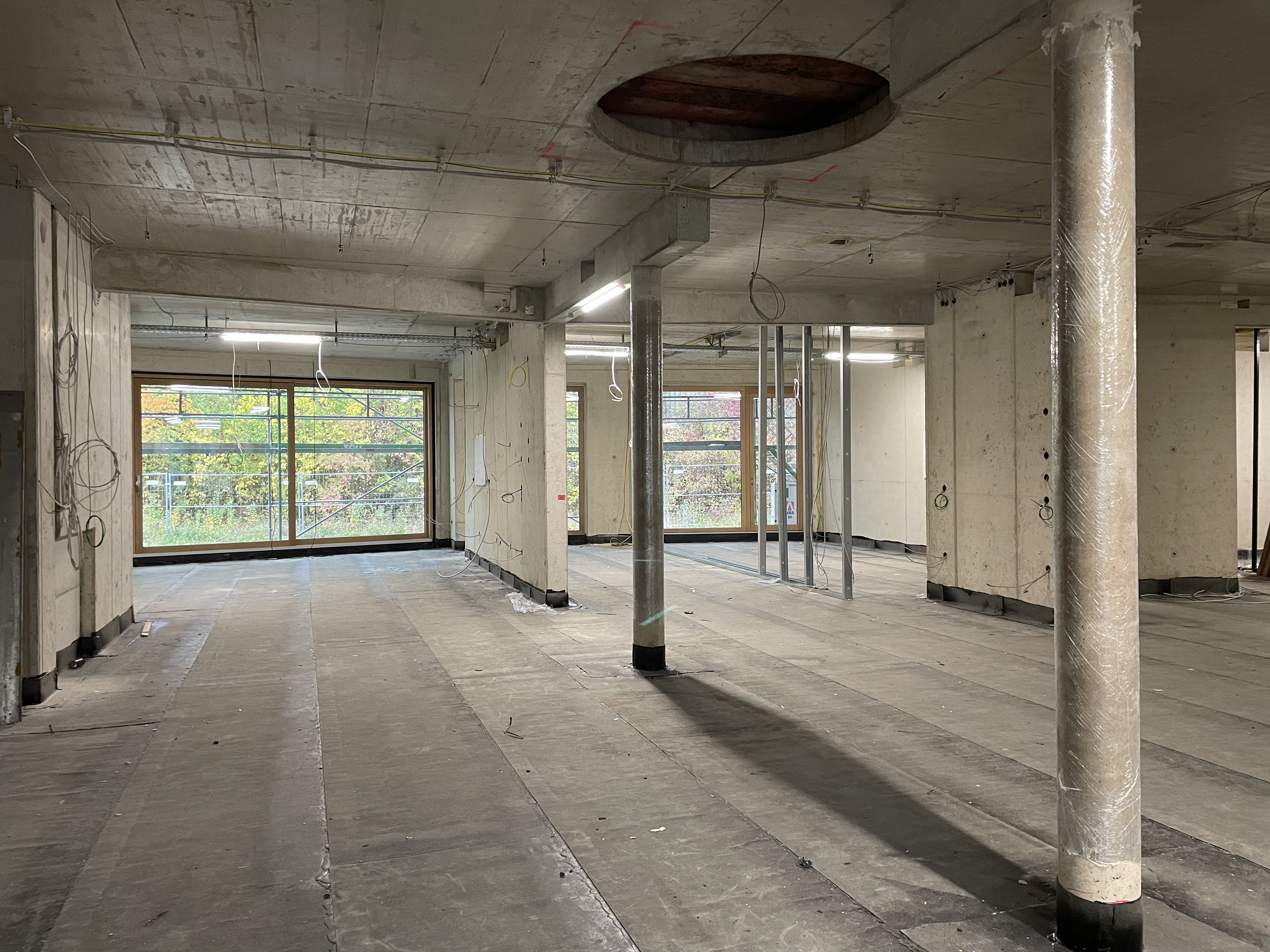Innenansicht im Erdgeschoss der Kita Merowingerstraße. Zwei Betonsäulen und betonierte Innenwände in einem Rohbau mit großen Fensterfronten. An der Betondecke sieht man eine große runde Aussparung. Mehrere Kabel hängen von der Decke.