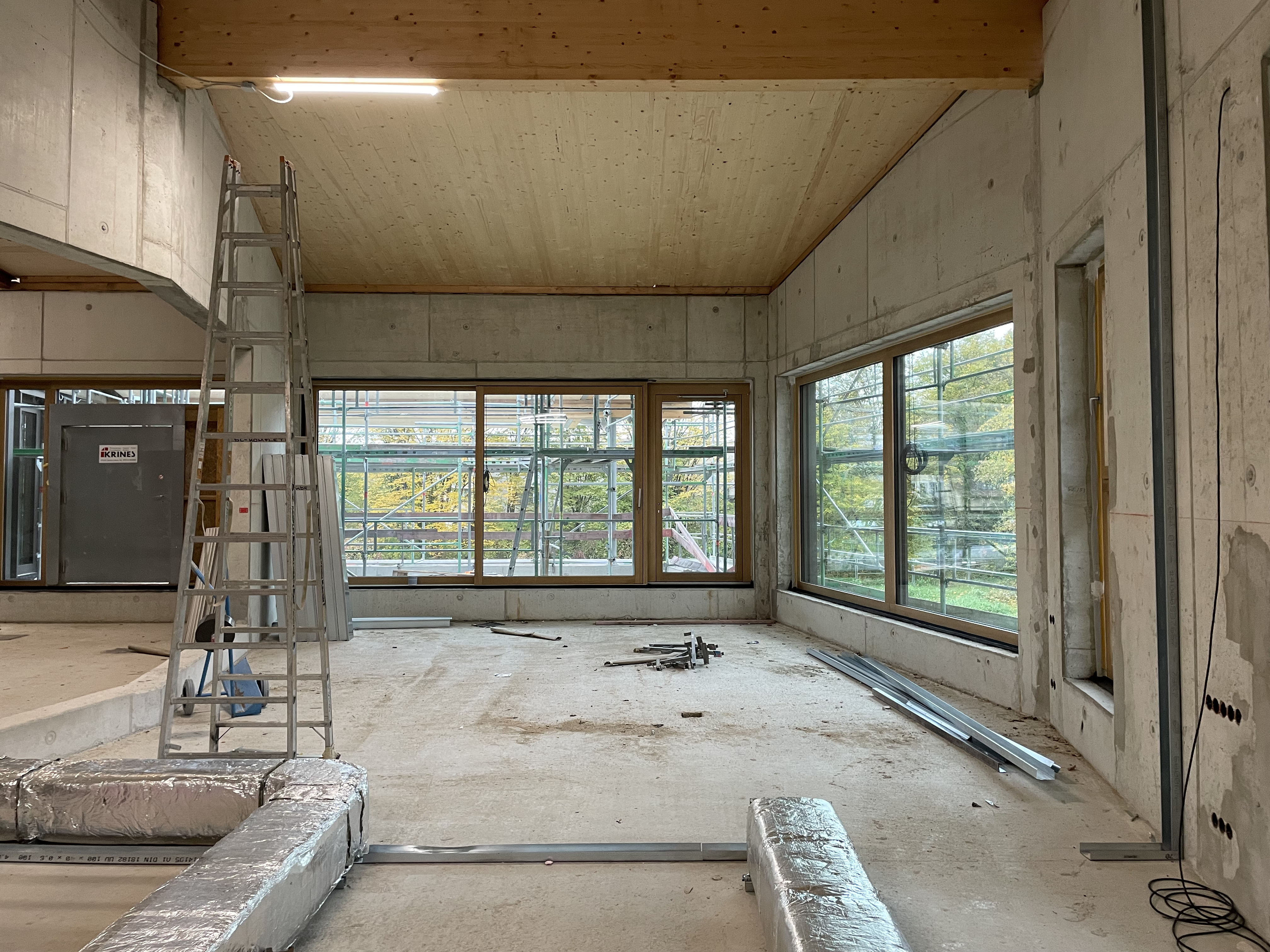 Einer der großzügigen Gruppenräume im Obergeschoss. Blick von innen auf die große Fensterfront in der Raumecke in einem Rohbau. Am Boden verlaufen isolierte große Rohre. Links im Bild befindet sich eine aufgestellte Leiter unter einer Lampe.