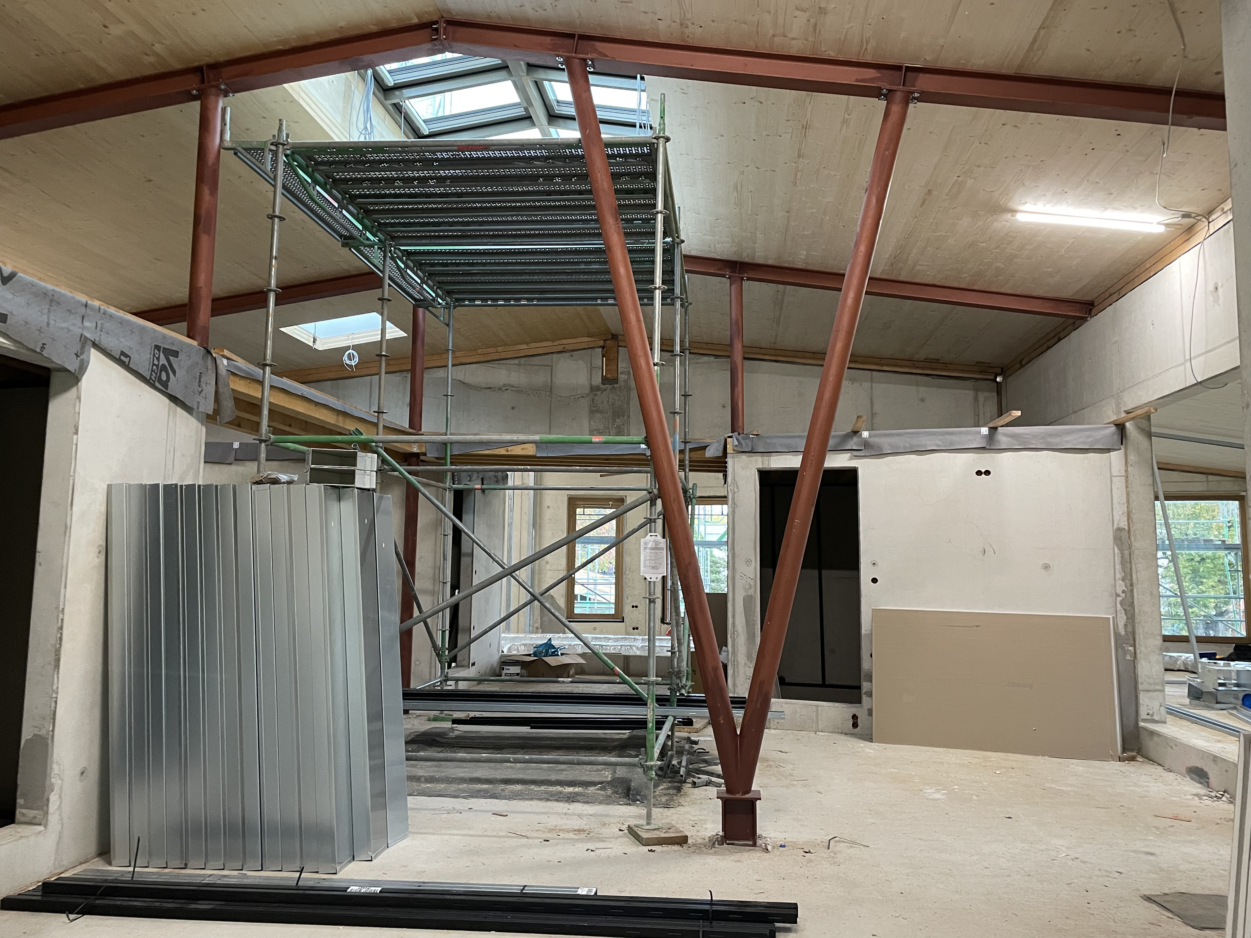 Stahlträger stützen das Dach mit einem großen Lichteinlass. Stahträger stützen ein von innen mit Holz verkleidetes Dach. Im Giebel befindet sich ein großes Dachflächenfenster, darunter ein Baugerüst. Man sieht betonierte Wände und die Abtrennung für kleinere Räume. Verschiedenes Baumaterial für den Innenausbau ist ein dem großen Raum verteilt.