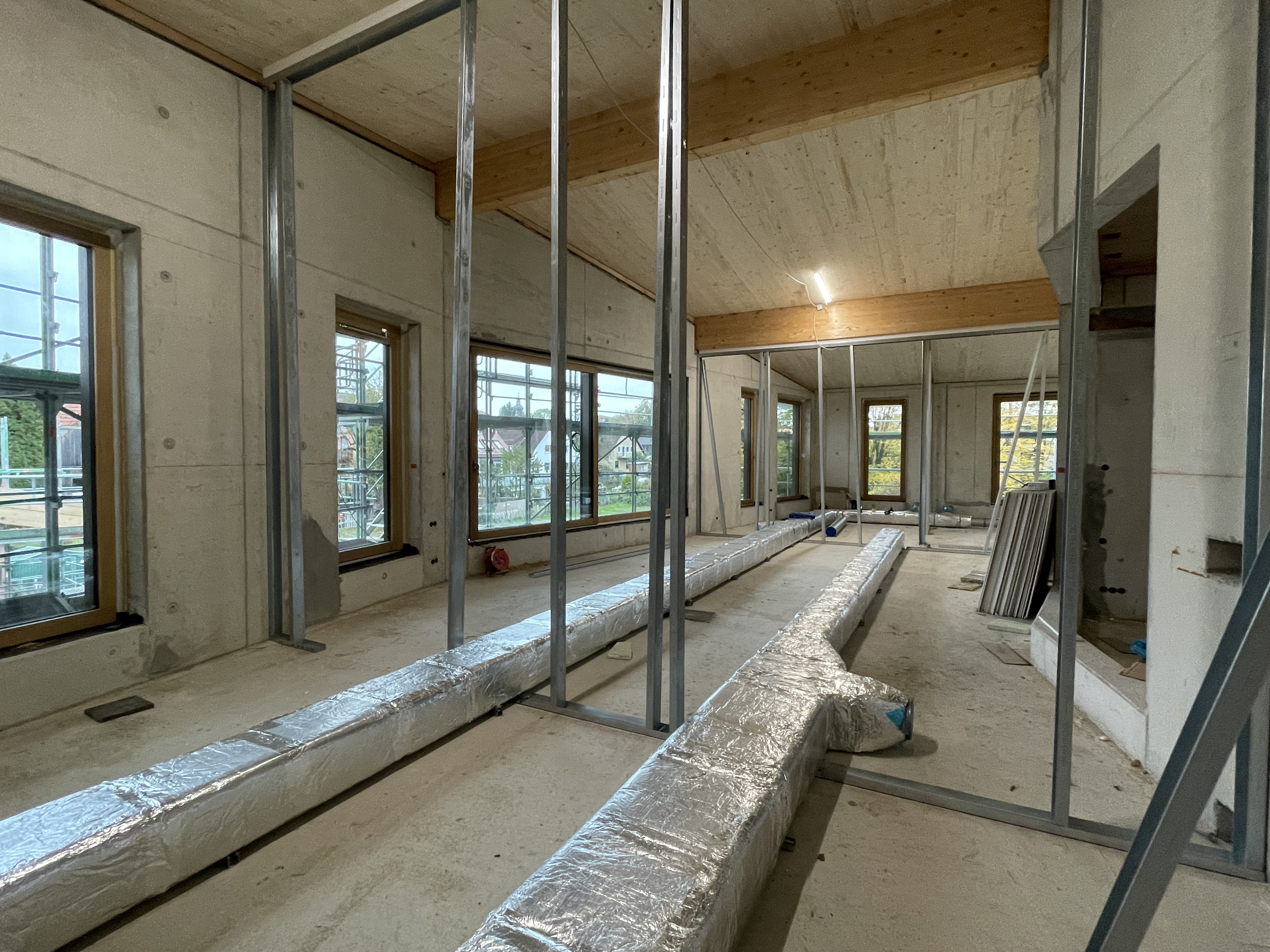 Mit dem Innenausbau und der Verlegung der Versorgungsleitungen und Rohre wurde bereits begonnen. Innenansicht eines Rohbaus mit Metallrahmen für den Innentrockenbau und zwei großen mit silberfarbigen Isoliermaterial umwickelten Versorgungsrohren.