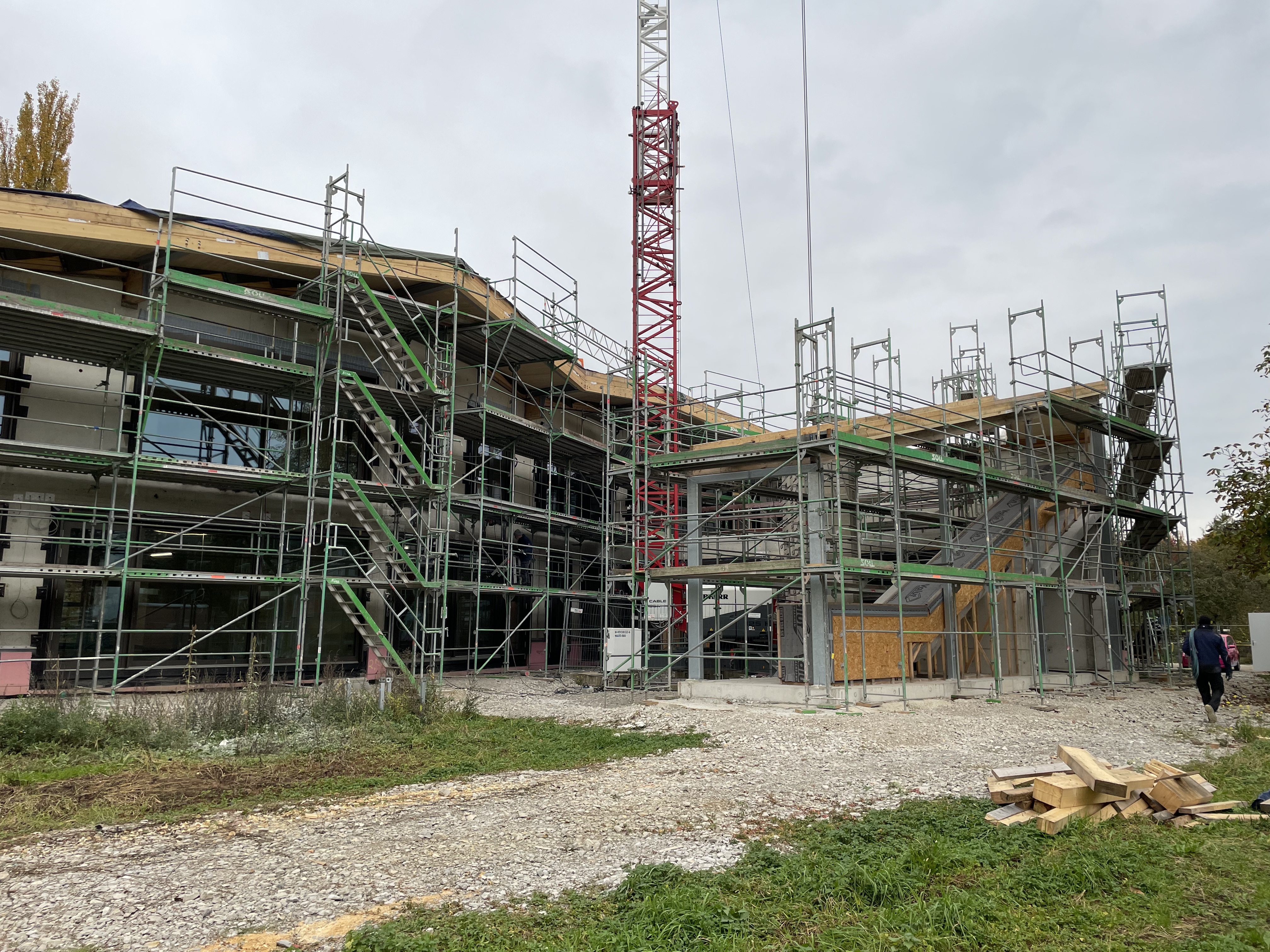 Die eingerüstete Außenfassade der Kita Merowingerstraße Der Rohbau der neuen Kita Merowingerstraße ist umgeben von Baustellengerüsten. In der Mitte steht ein Baukran. Im Vordergrund wechseln sich Wiesen- und Schotterflächen ab. Rechts vorne im Bild liegt ein Stapel mit Holzbaumaterial.