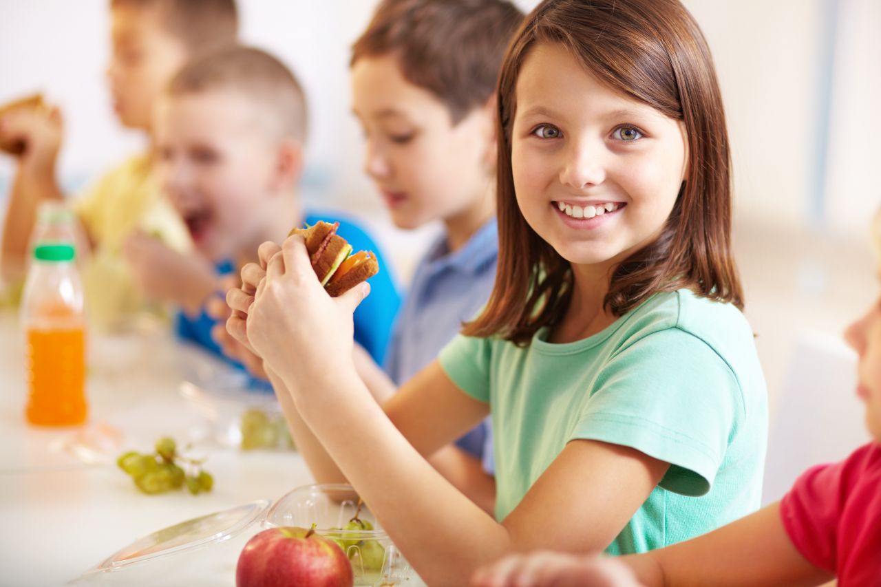 Mittagsbetreuung an Forchheimer Schulen Schulkinder sitzen während der Mittagsbetreuung an einem Tisch mit Getränken, Essen und Obst. Ein Mächen im Vordergrund lächelt in die Kamera.