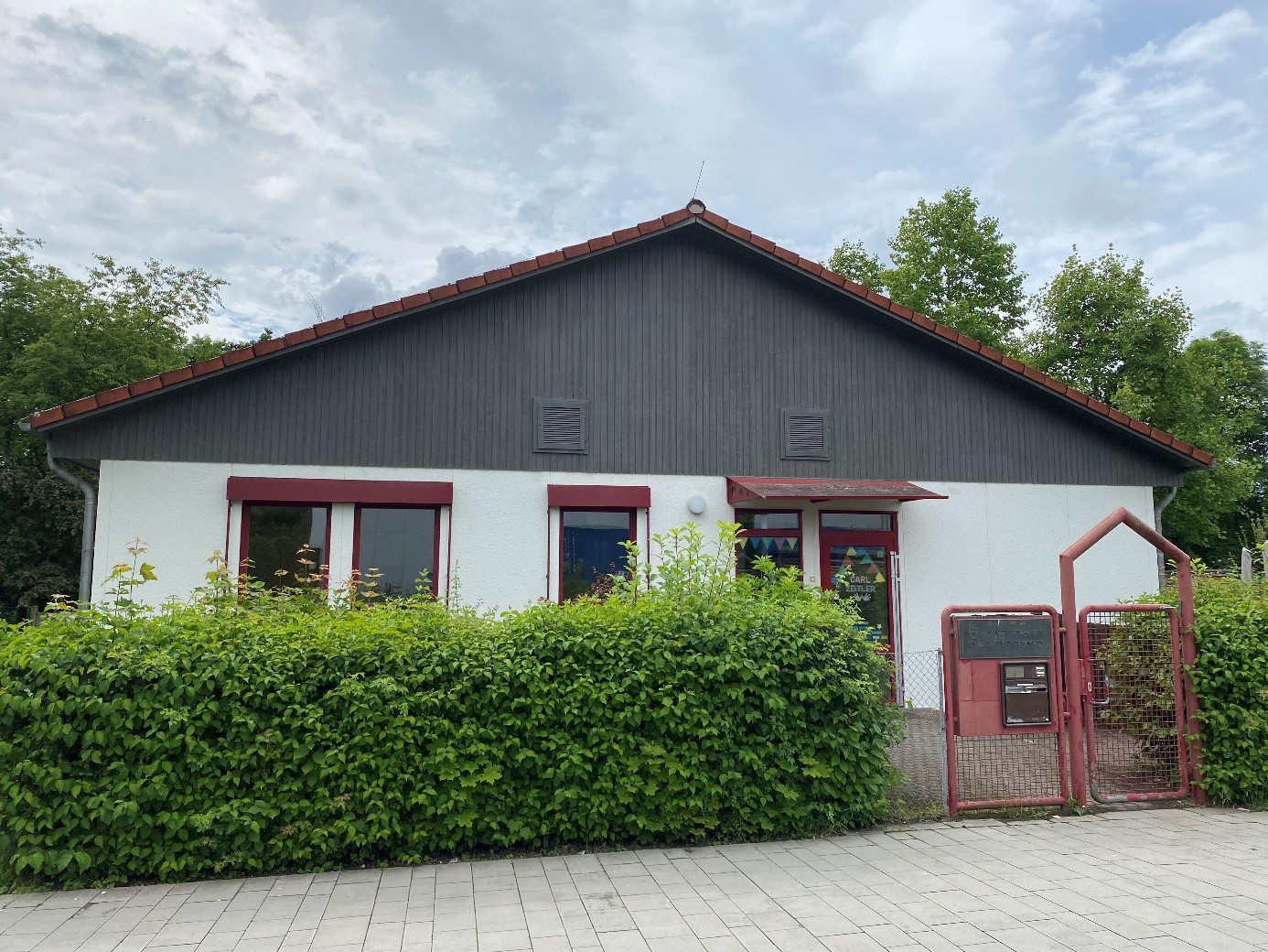 Eingangsbereich des Carl-Zeitler Kindergartens. Davor eine grüne Hecke und ein rotes Eingangstor