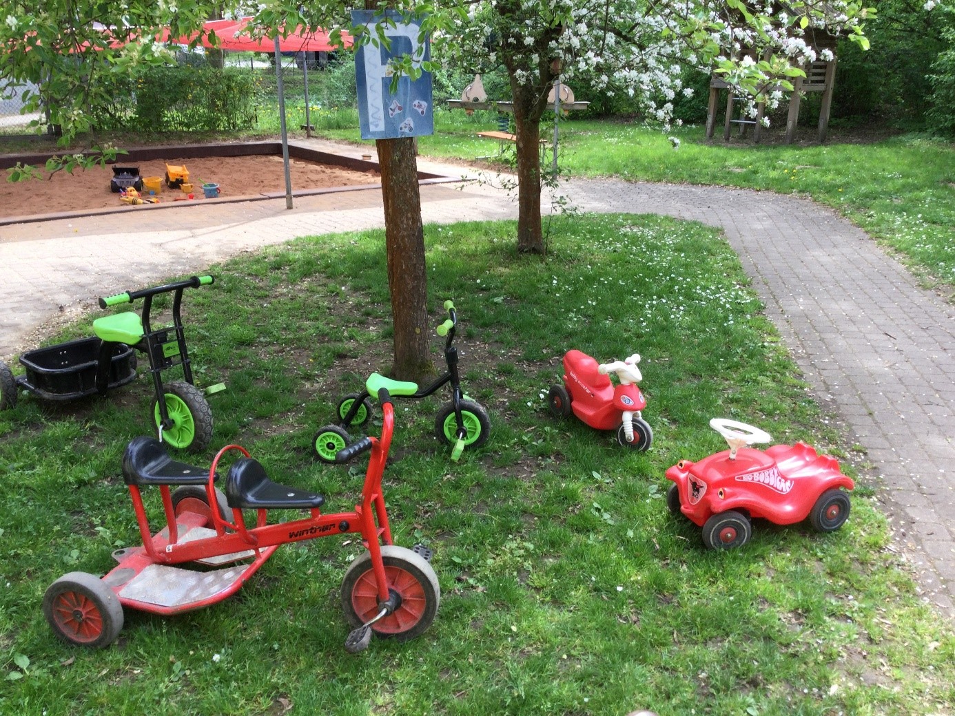 Mehrere Kinderräder und ein Bobbycar unter einem Baum auf eier Wiese.