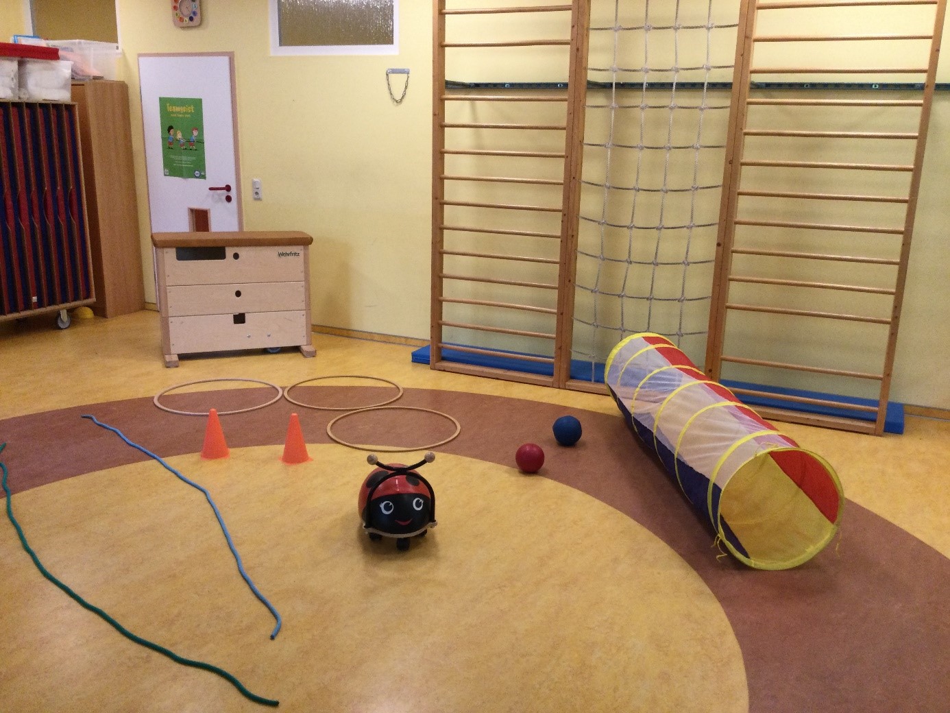 Ein Turmraum eines Kindergartens mit einer Sprossenwand, Seilen, Bällen, Reifen und einem bunten Kriechtunnel am Boden.