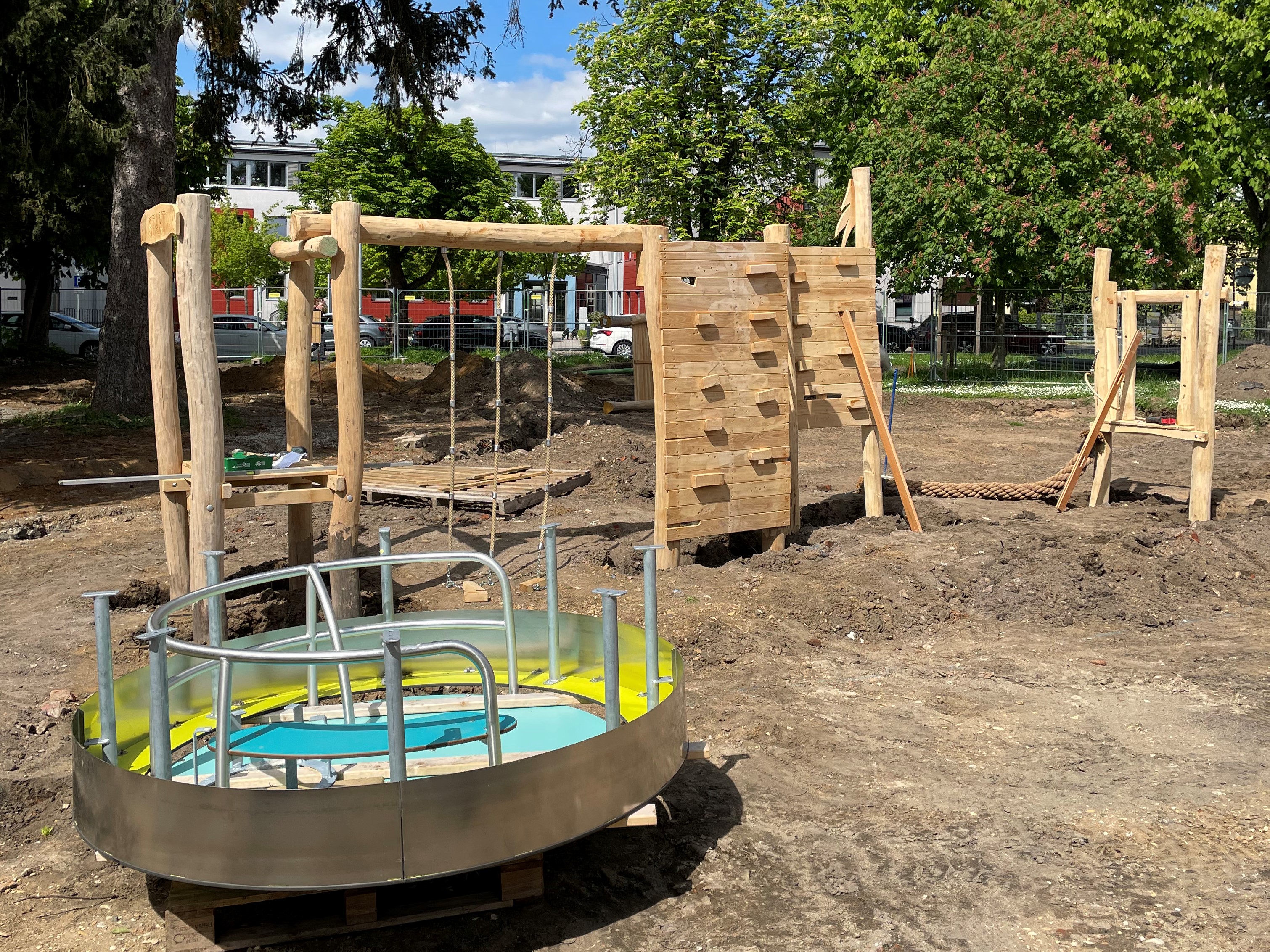 Aufbau von Spielgeräte mit grißem Holzanteil und einen Drehkarussel aus Metall auf einem Spielplatz.